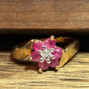 May include: A gold ring featuring a flower-shaped centrepiece composed of six round, vibrant pink rubies surrounding a small, central cluster of tiny, clear, sparkling diamonds. The band is crafted from polished yellow gold with a smooth finish.