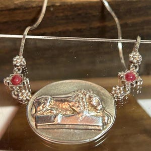 May include: A silver necklace featuring a large oval pendant with a detailed relief of a lion. The necklace has two decorative side pieces with red gemstones. The chain is a delicate snake chain style.