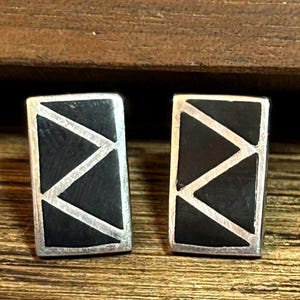 Könnte beinhalten: Ein Paar rechteckige Ohrstecker mit silbernem Rahmen und Einlage. Jeder Ohrstecker hat eine schwarze Steinoberfläche, die durch dünne, diagonale silberne Linien unterteilt ist und ein geometrisches Muster auf der Vorderseite bildet.