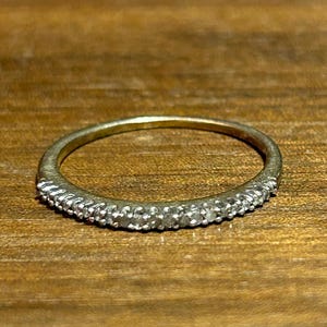 May include: A thin, gold-toned ring with a band of small, clear stones set along the top. The ring is displayed on a wooden surface, showcasing its delicate design and sparkle. The ring's simple elegance makes it a versatile accessory.