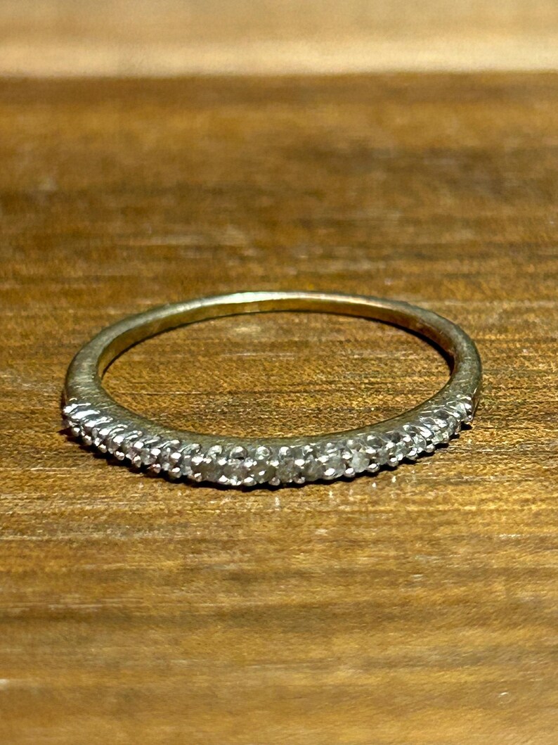May include: A thin, gold-toned ring with a band of small, clear stones set along the top. The ring is displayed on a wooden surface, showcasing its delicate design and sparkle. The ring's simple elegance makes it a versatile accessory.