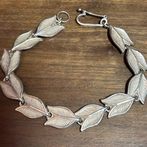 Puede incluir: Pulsera plateada con diseño de hojas. La pulsera está compuesta por múltiples eslabones en forma de hoja conectados por pequeños anillos. Las hojas tienen vetas detalladas y una superficie texturizada. El cierre es un simple cierre de gancho y ojo.