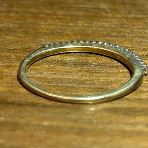 May include: A gold-colored ring with a band of small, clear stones set along the top. The ring is on a wooden surface, and the band is approximately 0.1 inches wide.