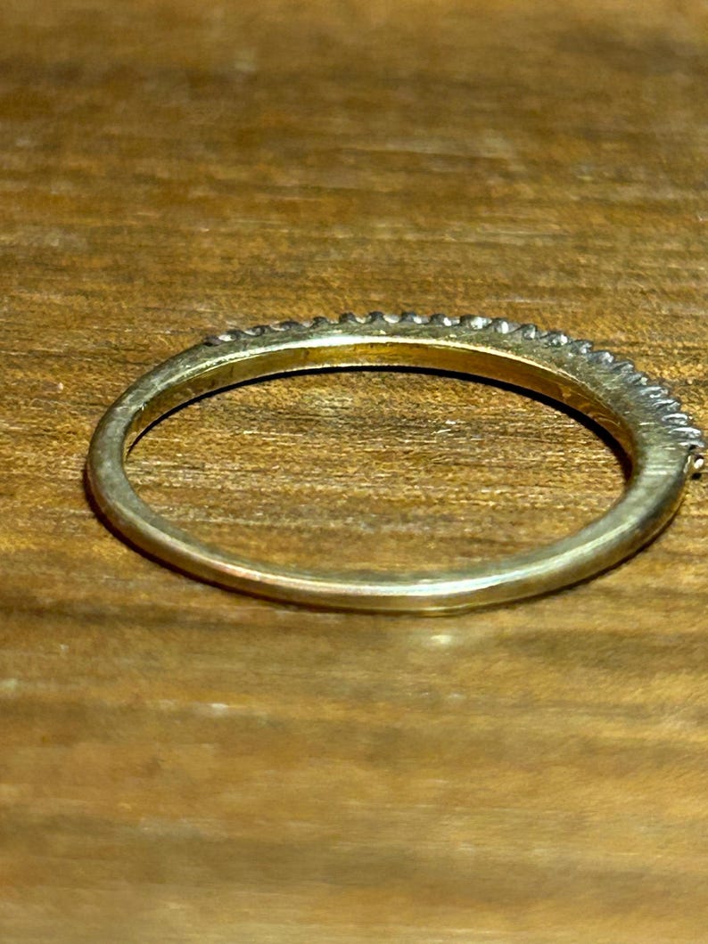 May include: A gold-colored ring with a band of small, clear stones set along the top. The ring is on a wooden surface, and the band is approximately 0.1 inches wide.