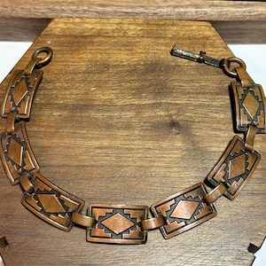 Puede incluir: Una pulsera de tono cobrizo con eslabones rectangulares, cada uno con un diseño en forma de diamante. La pulsera se exhibe sobre una superficie de madera, mostrando sus intrincados detalles y estética vintage. El cierre es de gancho y barra.