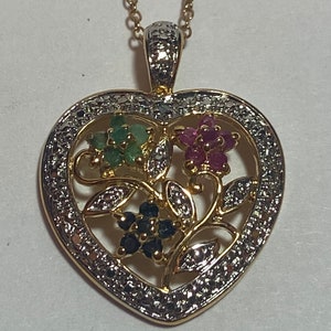 May include: A gold heart-shaped pendant necklace with a floral design featuring emerald, ruby, and sapphire gemstones. The pendant is surrounded by a border of small diamonds.