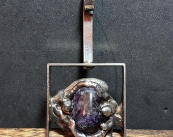 May include: A silver pendant featuring a rectangular frame with an abstract design. The pendant holds a large, oval-shaped, purple gemstone. The bail is a simple, rectangular bar. The pendant is displayed against a dark background.