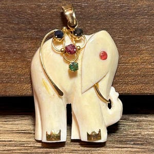 May include: Ivory-colored elephant pendant with gold accents. The pendant features a gold harness adorned with blue, red, and green gemstones. The elephant has a red gemstone eye and gold crown details on its feet.