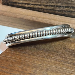 May include: A silver cuff bracelet featuring a central twisted rope wire design flanked by two smooth, polished silver bands. The metal has a reflective finish with subtle dark oxidation in the crevices of the twisted wire detail.