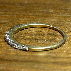 May include: A gold-toned ring with a band of small, clear gemstones set along the top half. The ring is set against a wooden surface, showcasing its delicate design and sparkle.