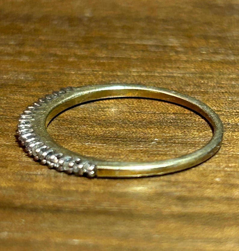 May include: A gold-toned ring with a band of small, clear gemstones set along the top half. The ring is set against a wooden surface, showcasing its delicate design and sparkle.
