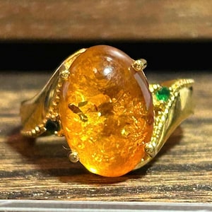 May include: A gold ring featuring a large, oval amber stone with inclusions. The ring has a detailed gold band with small green gemstones on the sides. The amber is a warm, translucent orange color.