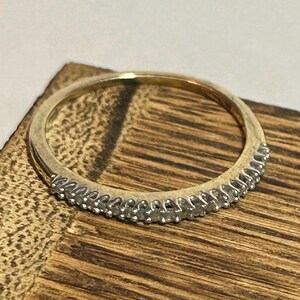 May include: A gold band ring with a row of small, clear gemstones set along the top. The ring is set against a wooden surface, showcasing its design and sparkle. The ring is a delicate piece of jewelry.
