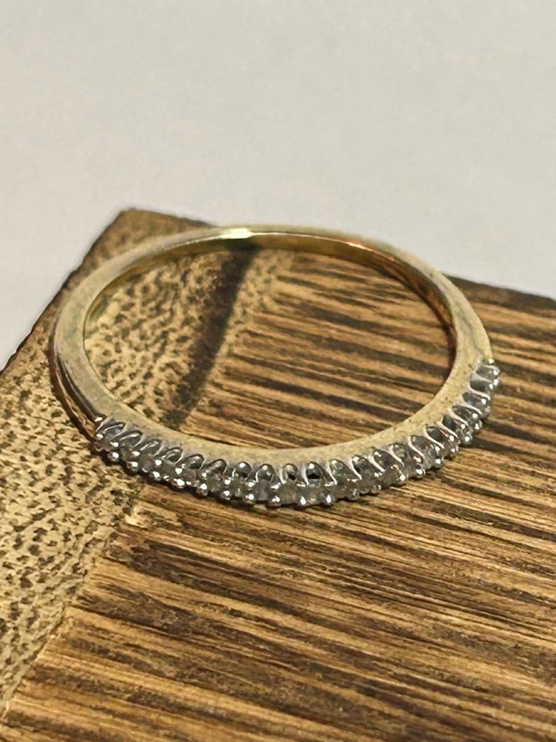 May include: A gold band ring with a row of small, clear gemstones set along the top. The ring is set against a wooden surface, showcasing its design and sparkle. The ring is a delicate piece of jewelry.