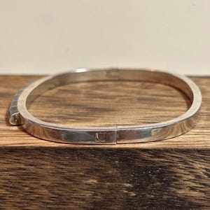 May include: A silver-colored bangle bracelet with a rectangular clasp. The bracelet has a smooth, polished surface and a simple, minimalist design. The bracelet is shown on a wooden surface.