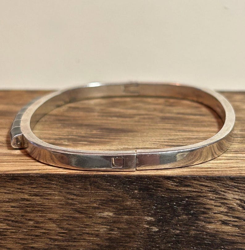 May include: A silver-colored bangle bracelet with a rectangular clasp. The bracelet has a smooth, polished surface and a simple, minimalist design. The bracelet is shown on a wooden surface.
