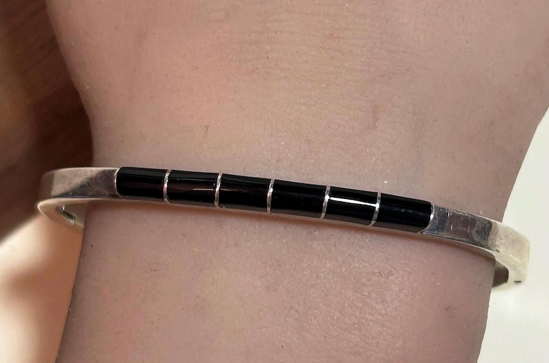 May include: A silver cuff bracelet with a series of six rectangular black onyx stones. The bracelet is worn on a wrist, showcasing the contrast between the shiny metal and the dark stones. A simple, elegant piece of jewelry.