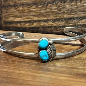 Puede incluir: Una pulsera de plata tipo brazalete con dos piedras turquesas ovaladas dispuestas verticalmente en el centro. La pulsera presenta un diseño de doble banda y un elemento decorativo de hoja de plata junto a las piedras.
