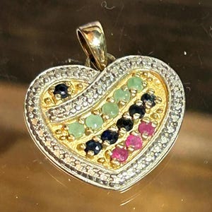 May include: A heart-shaped pendant with a gold bail and a gold-colored interior. The pendant is encrusted with small, colorful gemstones, including green, blue, and pink stones, and is surrounded by a border of small, clear stones.