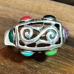 May include: A silver ring featuring a rectangular openwork frame with a central double-spiral design. The ring is accented with small, smooth, cabochon-style stones in various colors, including red, black, purple, light green, turquoise, and dark green.