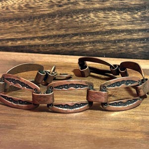 Puede incluir: Una pulsera de eslabones de cobre con segmentos ovalados que presentan patrones grabados oscuros y hundidos. El metal tiene un acabado metálico cálido y el diseño consiste en motivos geométricos repetidos unidos por pequeñas anillas de cobre.