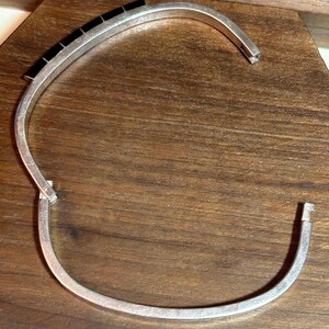 May include: A silver-toned cuff bracelet with a row of black rectangular stones. The bracelet is open, revealing the clasp and the ends of the band. The jewelry is displayed on a wooden surface.