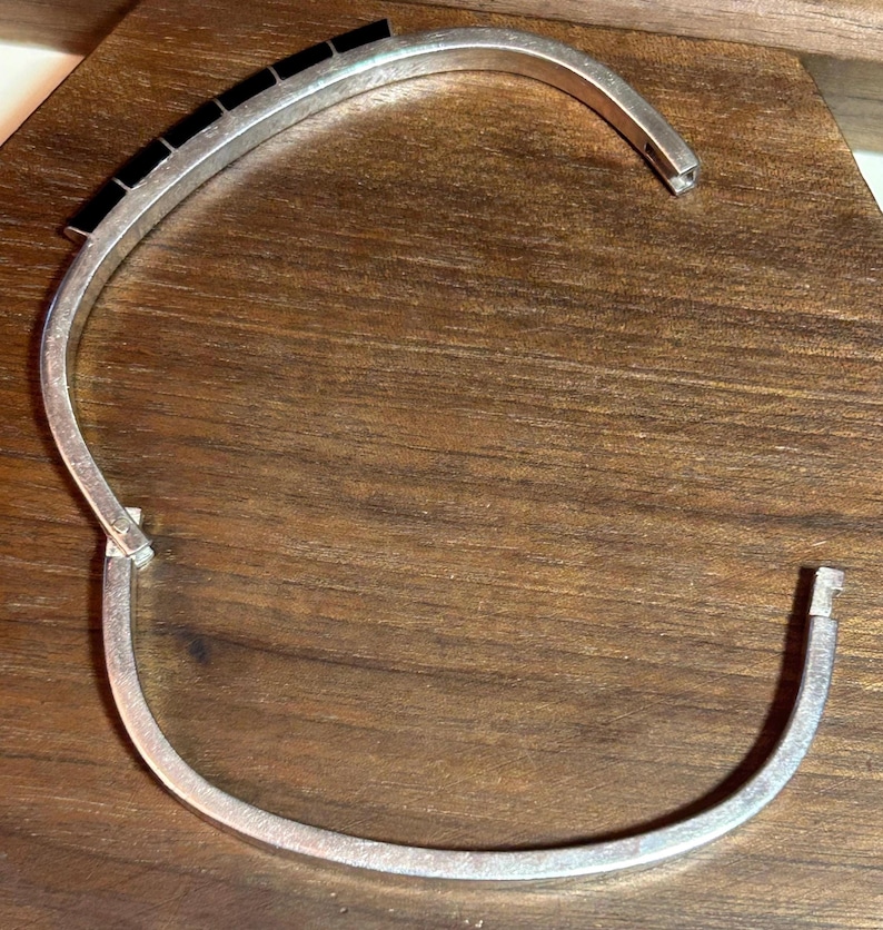 May include: A silver-toned cuff bracelet with a row of black rectangular stones. The bracelet is open, revealing the clasp and the ends of the band. The jewelry is displayed on a wooden surface.