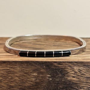 May include: Silver-toned bangle bracelet with a rectangular shape. The bracelet features a series of five black rectangular inlays. The bracelet is displayed on a wooden surface.