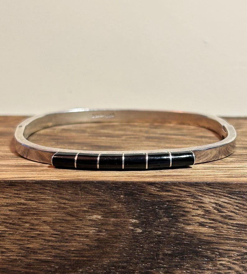 May include: Silver-toned bangle bracelet with a rectangular shape. The bracelet features a series of five black rectangular inlays. The bracelet is displayed on a wooden surface.