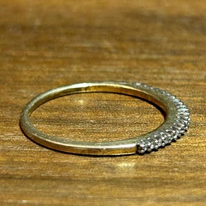 May include: A gold-toned ring with a band of small, clear stones set along the top half. The ring is set against a warm, wooden surface, highlighting its metallic sheen and the sparkle of the stones. The ring is a piece of jewelry.