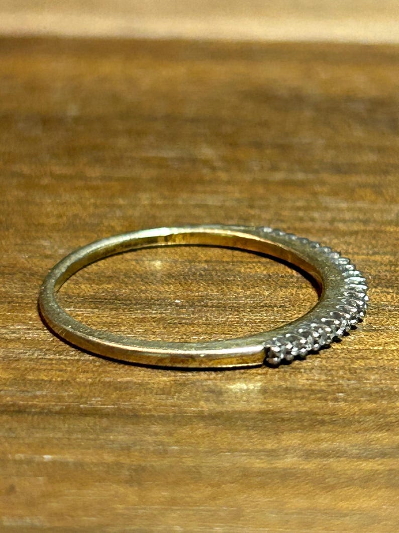 May include: A gold-toned ring with a band of small, clear stones set along the top half. The ring is set against a warm, wooden surface, highlighting its metallic sheen and the sparkle of the stones. The ring is a piece of jewelry.