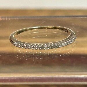 May include: A thin gold band ring featuring a row of small, clear, round-cut stones set along the front half of the band. The metal has a polished finish, and the stones are arranged in a continuous line, creating a sparkling effect across the surface.