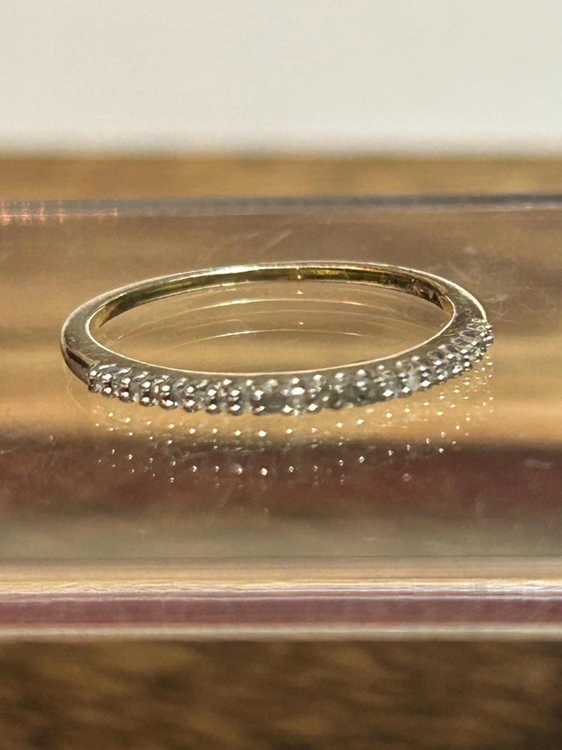 May include: A thin gold band ring featuring a row of small, clear, round-cut stones set along the front half of the band. The metal has a polished finish, and the stones are arranged in a continuous line, creating a sparkling effect across the surface.
