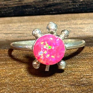 May include: A silver ring featuring a turtle design. The ring has a bright pink, round opal stone as the turtle's shell, with four silver legs. The ring is set on a wooden surface.