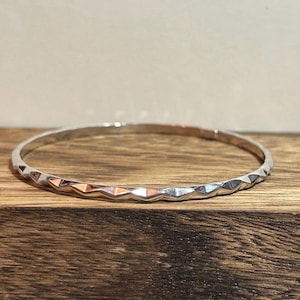 May include: A silver bangle bracelet with a geometric, faceted design. The bracelet is circular and has a textured surface, reflecting light. The bangle is displayed on a wooden surface.