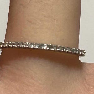 May include: A delicate silver ring with a row of small, sparkling diamonds. The ring is set on a finger, showcasing its elegant design and subtle brilliance. The diamonds are evenly spaced, creating a continuous line of light.