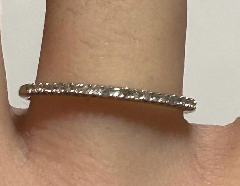 May include: A delicate silver ring with a row of small, sparkling diamonds. The ring is set on a finger, showcasing its elegant design and subtle brilliance. The diamonds are evenly spaced, creating a continuous line of light.