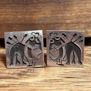 May include: A pair of square silver-toned cufflinks. Each cufflink features a stylized design of a figure with a hat and arrows. The background is a dark wood grain.