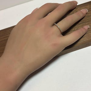 May include: A gold-colored ring with small, clear stones set along the band. The ring is on a person's finger, resting on a wooden surface. The ring appears to be a wedding band or a similar type of jewelry.