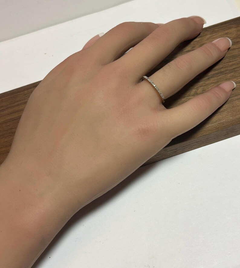 May include: A gold-colored ring with small, clear stones set along the band. The ring is on a person's finger, resting on a wooden surface. The ring appears to be a wedding band or a similar type of jewelry.