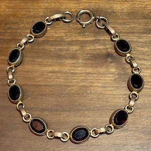 May include: A gold-toned bracelet featuring oval-shaped, dark red gemstones. The bracelet has a delicate chain design with a clasp closure. The gemstones are set in a gold-colored metal, creating a vintage aesthetic.
