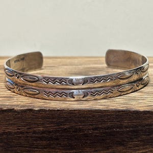 Pode incluir: Duas pulseiras de prata esterlina com desenhos gravados intrincados. Cada pulseira apresenta um padrão repetitivo de formas ovais, ziguezagues e motivos estilizados. A palavra "STERLING" está estampada no interior de uma pulseira. As pulseiras são exibidas em uma superfície de madeira.