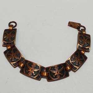 Pode incluir: Um bracelete de cobre com um padrão repetido de figuras estilizadas. As figuras são de cor castanho escuro e estão sobre um fundo de cobre.