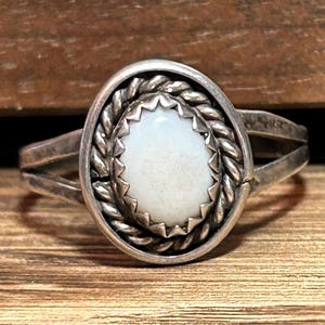 May include: Oval-shaped silver ring with a light-colored stone. The stone is surrounded by a decorative border and a braided silver band. The ring has a split shank design.