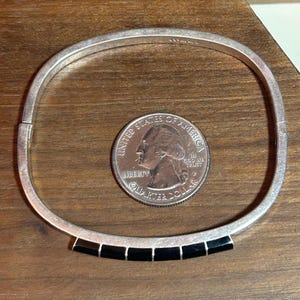 May include: A silver-toned hinged bangle bracelet with a row of six black rectangular insets. A United States quarter coin is next to the bracelet for size comparison. The bracelet is on a wooden surface.
