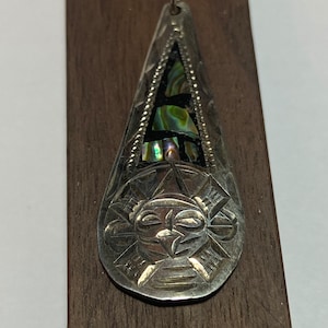 May include: A silver pendant with a geometric design and a piece of abalone shell in the center. The pendant is in the shape of a teardrop and has a loop at the top for hanging.