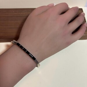May include: A silver bracelet with a series of rectangular black inlays. The bracelet is worn on a wrist, showcasing its design and craftsmanship. The background is a wooden surface.