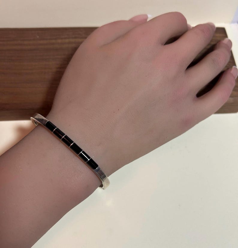 May include: A silver bracelet with a series of rectangular black inlays. The bracelet is worn on a wrist, showcasing its design and craftsmanship. The background is a wooden surface.