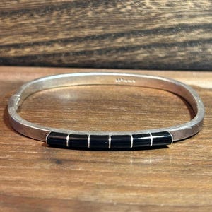 May include: A silver bracelet with a row of six black rectangular inlays. The bracelet is set against a wooden surface. The bracelet is a piece of jewelry.