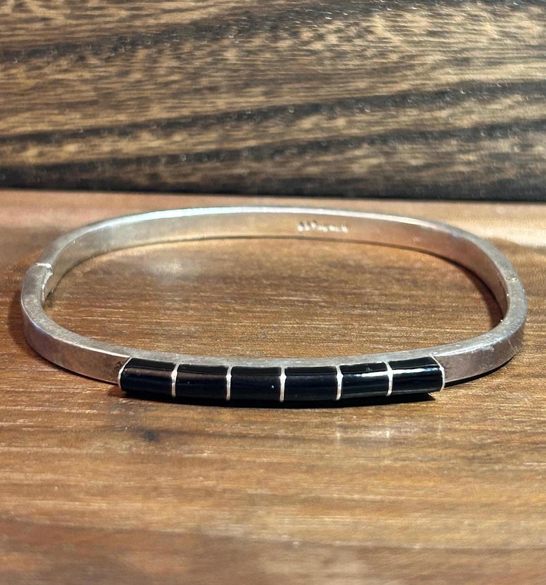 May include: A silver bracelet with a row of six black rectangular inlays. The bracelet is set against a wooden surface. The bracelet is a piece of jewelry.